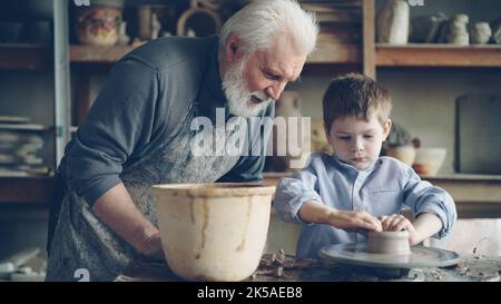 Konzentrierter Junge formt Ton in einen Keramiktopf auf dem drehenden Wurfrad und sein liebevoller erfahrener Großvater spricht mit ihm. Töpferei und Familientradition Konzept. Stockfoto