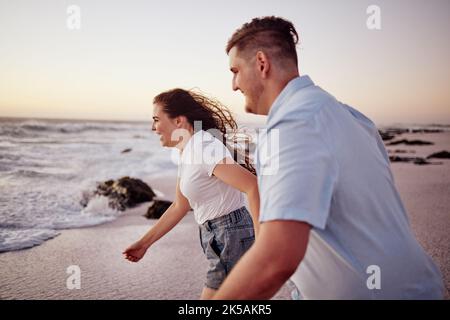 Liebe, Strand und Paar halten sich die Hände, die an einem romantischen Sonnenuntergang-Abend draußen in der Natur zum Wasser laufen. Lächeln, Romantik und glückliche Frau auf Stockfoto