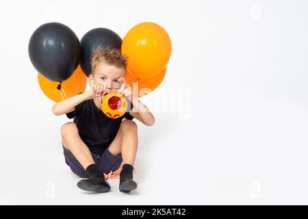 Halloween Für Kinder. Ein Junge, der einen leeren Eimer in Form eines Kürbisses zeigt und schwarze und orangefarbene Ballons hält und Süßigkeiten auf einem weißen Studiohintergrund isst Stockfoto