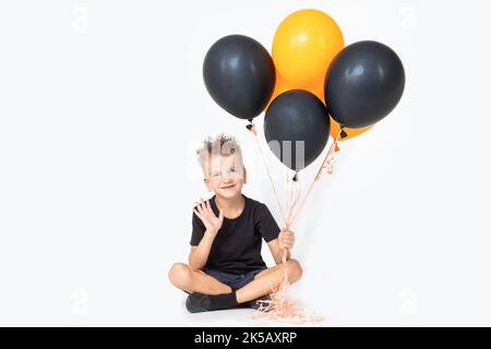 Ein fröhlicher, fröhlicher Junge in einem Zaubererhut begrüßt eine winkende Hand, die schwarze und orangefarbene Ballons hält und auf einem weißen Studiohintergrund sitzt. Hallo Halloween Stockfoto