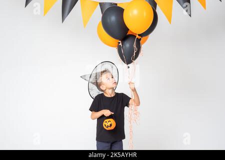 Ein fröhlicher, fröhlicher Halloween-Junge in einem Zaubererhut mit einem Kürbis-förmigen Eimer und schwarzen und orangen Ballons auf einem weißen Studiohintergrund mit einem Ga Stockfoto