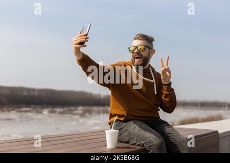 Bärtiger Mann genießt den Frühling und nimmt Selfie-Fotos auf der Promenade mit dem Smartphone auf. Technologien Freizeit und gen z Konzept Stockfoto