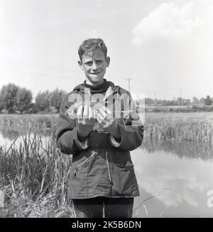 1960s, historisch, ein Teenager-Junge mit Kapuzenoberteil, Seitentaschen und Kordelzug am Bund, der an einem Flussufer steht und seinen Fang zeigt, England, Großbritannien. Die Jugend nahm an einem Fischfang Teil, Stockfoto