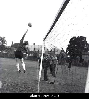 1960s, historisch, draußen auf einem Feld, ein Amateurfußball, ein Torwart, der einen Spar macht, sich ausdehnt, um den Ball über die Querlatte zu schieben, England, Großbritannien, beobachtet von einigen Zuschauern, die in der Nähe der Torpfosten stehen. Stockfoto