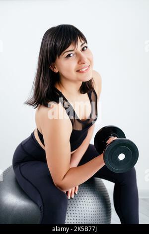 Starke Körperkondition. Athletische Frau. Fitnessstudio für Krafttraining. Heimtraining. Glückliche Dame in schwarzer Sportbekleidung macht Hanteln Training sitzende fit Ball in weißem Backg Stockfoto