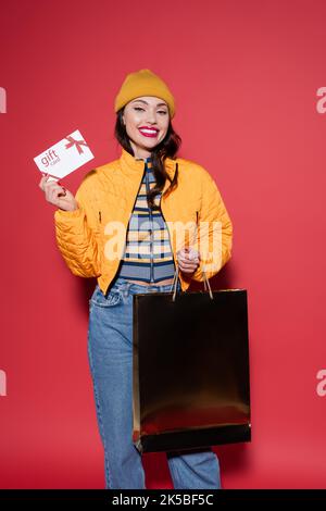 Glückliche junge Frau in orange Mütze Hut mit Geschenkkarte und Einkaufstasche auf rot, Stock Bild Stockfoto