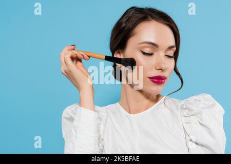 Brunette junge Frau in weißer Bluse hält kosmetischen Pinsel in der Nähe Wange isoliert auf blau, Stock Bild Stockfoto