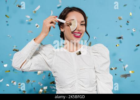 Fröhliche junge Frau in weißer Bluse, die Augen mit kosmetischen Pinsel in der Nähe fallenden Konfetti auf blau bedeckt, Stockbild Stockfoto