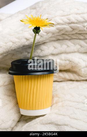 Heiß-aromatischer Kaffee in einer gelben Tasse zum Mitnehmen und die Blume des Dandelions für die Morgenromantik steht auf einem warmen Strickpullover. Konzept des kalten Herbstes und Treffen der Liebhaber. Vertikales Foto. Speicherplatz Kopieren Stockfoto