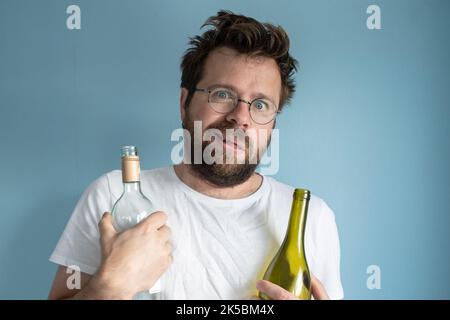 Der zaghafte, bärtige Mann umarmt zwei leere Weinflaschen, die er trank, er fühlt sich schlecht und leidet an einem Kater. Konzept der Alkoholabhängigkeit. Stockfoto