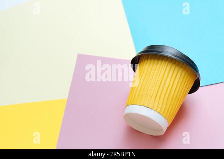 Die gelbe Kaffeetasse zum Mitnehmen liegt in kreativem Chaos auf farbigem Papier auf dem Tisch des Designers. Blauer, orangefarbener und rosafarbener Hintergrund. Platz für Text kopieren Stockfoto