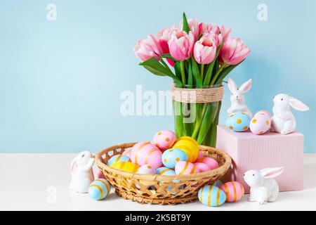 Ostern und Frühlingsferien Grußkartenkonzept. Rosa Tulpen, osterhasen mit bunten Eiern im Korb und blauem Hintergrund. Bemalte Ostereier Stockfoto