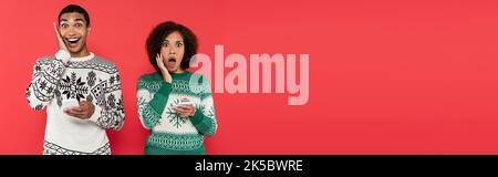 Schockierte afroamerikanische Frau in Winterpullover mit Handys und Blick auf Kamera isoliert auf roten, Banner, Stock Bild Stockfoto