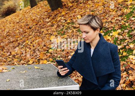 Die kurzhaarige Blondine setzt im Herbst ein Smartphone ein, das an einem Tisch im Park sitzt. Stockfoto