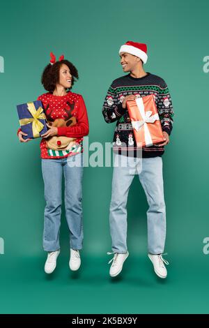 Fröhliches afroamerikanisches Paar in Pullovern mit weihnachtlichen Mustern schweben mit Geschenkschachteln auf grünem Hintergrund, Stockbild Stockfoto
