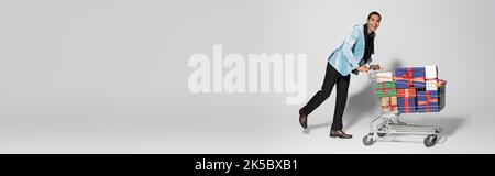 In voller Länge von eleganten afroamerikanischen Mann bewegt Warenkorb mit Geschenk-Boxen auf grauem Hintergrund, Banner, Stock Bild Stockfoto