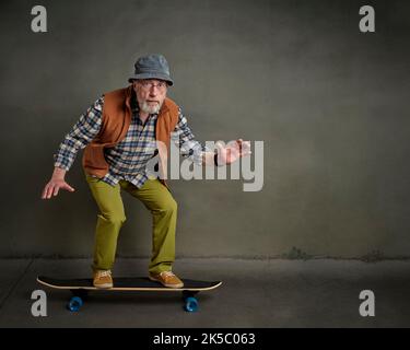 Bärtiger älterer Mann mit runder Brille und einem Eimer Hut reitet auf einem langen Skateboard in einer grunge städtischen Umgebung Stockfoto