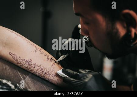 Portrait des Mannes Tattoo Meister mit Dreadlocks zeigt Prozess der Schöpfung Tattoo auf weiblichen Körper unter Lampe Licht. Professioneller Künstler, der im Salon arbeitet Stockfoto