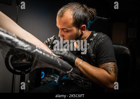 Portrait des Mannes Tattoo Meister mit Dreadlocks zeigt Prozess der Schöpfung Tattoo auf weiblichen Körper unter Lampe Licht. Professioneller Künstler, der im Salon arbeitet Stockfoto