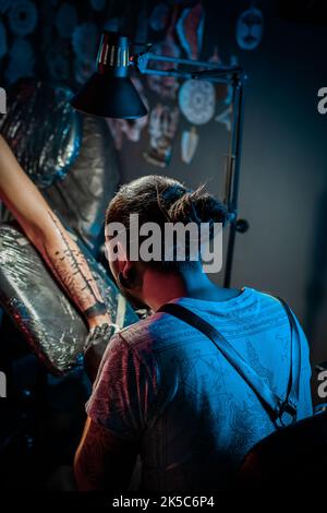 Portrait des Mannes Tattoo Meister mit Dreadlocks zeigt Prozess der Schöpfung Tattoo auf weiblichen Körper unter Lampe Licht. Professioneller Künstler, der im Salon arbeitet Stockfoto