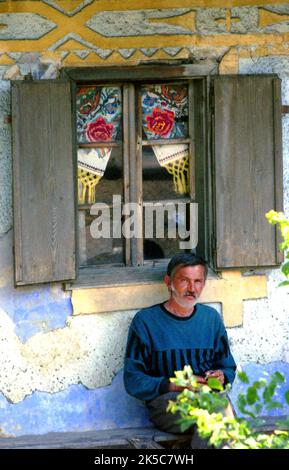 Bistrița-Năsăud County, Rumänien, 2000. Mann, der auf einer Bank außerhalb der farbenfrohen Fassade seines ursprünglichen Hauses sitzt. Stockfoto