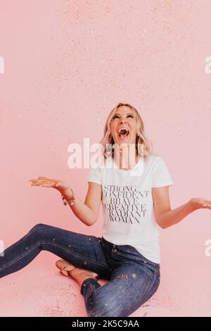 Frau sitzt auf dem Boden und lacht, nachdem sie Gold-Glitzer geworfen hat Stockfoto