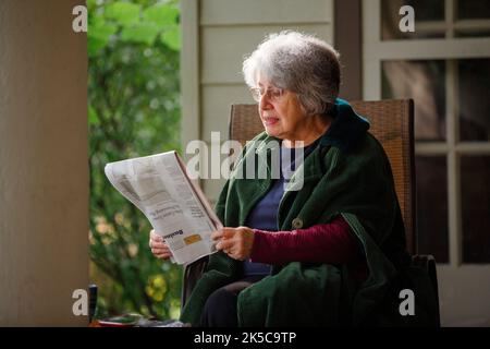 Eine ältere Frau im warmen Mantel sitzt auf der Veranda und liest Zeitung Stockfoto