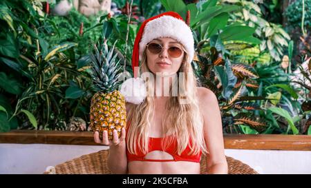 Schöne lächelnde Blondine im Weihnachtsmann-Hut, die in die Kamera blickt und Ananas mit Sonnenbrille vor dem Hintergrund grüner tropischer Pflanzen hält. Konzept Stockfoto