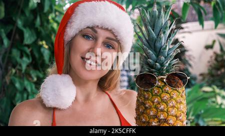 Schöne lächelnde Blondine im Weihnachtsmann-Hut, die in die Kamera blickt und Ananas mit Sonnenbrille vor dem Hintergrund grüner tropischer Pflanzen hält. Konzept Stockfoto