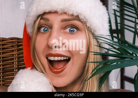 Lustige blonde Frau mit breitem Lächeln und Weihnachtsmütze, die mit überraschten Augen in die Kamera schaut. Mädchen feiert Weihnachten in den Tropen. Festliche Stimmung Stockfoto