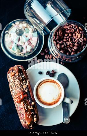 Ansicht des traditionellen französischen Dessertes eclair mit dunkler Schokolade und Haselnüssen auf dem Tisch im Café mit einer Tasse Kaffee, Kaffeebohnen und Mini-Marschmalow Stockfoto