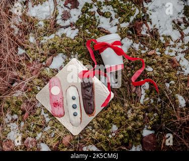 Draufsicht auf ein traditionelles französisches Dessert eclair auf goldenem Hintergrund im Wald mit Moos und einer Papierkaffeetasse mit rotem Band. Geschenkkonzept Stockfoto