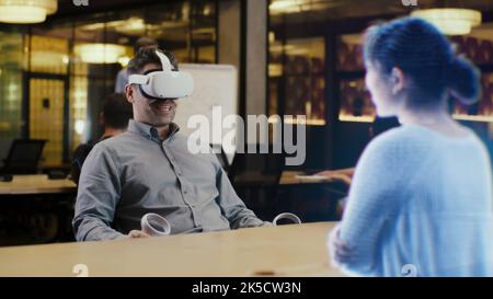 Ein Mann mit Virtual-Reality-Headset sitzt mithilfe von Joysticks auf einem Bürotisch. Führt ein Interview mit einem Kandidaten durch, der das Augmented Reality 3D-Modellkonzept verwendet. Hologramm des Mädchens erscheint vor dem Mann. Stockfoto