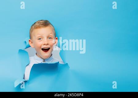 Happy cute boy hat Spaß auf blauem Hintergrund Wand gespielt, klettert durch ein Loch in das Papier. Helle und lustige Emotionen des Jungen Stockfoto