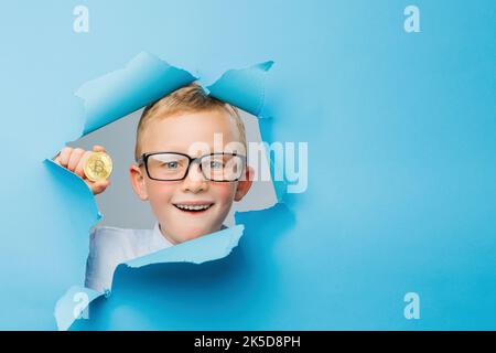 Happy cute Businessboy in Brillen hat Spaß und hält Bitcoin in der Hand auf blauem Hintergrund Wand, klettert durch ein Loch in das Papier. Hell und Stockfoto