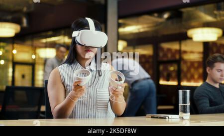 Asiatische Büroangestellte, die ein VR-Headset und drahtlose Controller verwendet und Daten in der virtuellen Realität sieht. Arbeiten Sie im modernen Büro eines Hightech-Unternehmens mit verschiedenen Kollegen. Digitale Cyberspace-Technologie Stockfoto