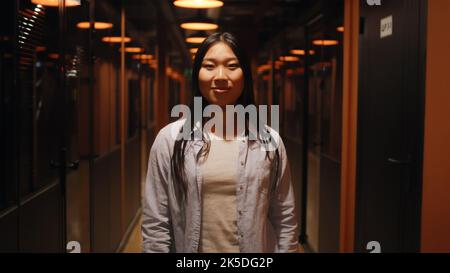 Glückliche und erfolgreiche asiatische Frau, die im Büroflur steht, lächelt und die Kamera anschaut. Motivierte Büroangestellte im Korridor des modernen Geschäftszentrums der Marketingfirma Stockfoto