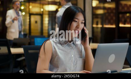Asiatische Frau, die mit dem Kunden telefoniert, während sie in einem modernen Büro in der Nähe von Kollegen arbeitet, mit einem Laptop und einem digitalen Tablet. Geschäftsfrau diskutiert über E-Commerce-Startup. Strategie des Unternehmens Stockfoto