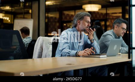 Reife Büroangestellte in Brillen, die telefonieren, Audionachrichten aufzeichnen und in einem modernen Business Center in der Nähe von Mitarbeitern auf einem Laptop arbeiten. E-Commerce-Marketing-Unternehmen Stockfoto