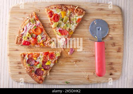 Pizza mit Salami, Pfefferoni, Tomaten und Zwiebeln auf einem Tisch und ein Pizzaschneider daneben - Blick von oben Stockfoto