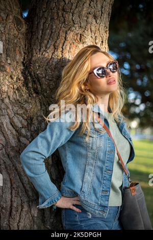 Lächelnde, stilvolle junge blonde Frau, die sich in der Natur an einen Baum lehnt Stockfoto