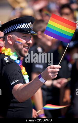Brighton & Hove Pride Festival, Brighton & Hove, East Sussex, England. Britischer Polizist in Uniform mit Regenbogenfahne auf dem Gesicht gemalt und mit der Hand gewellt Stockfoto