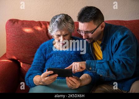 Oma hält ein Smartphone mit einem Videoclip, während ihr Enkel im Wohnzimmer sitzt. Zwei Generationen online mit dem Telefon auf der Couch zu Hause. Elderl Stockfoto