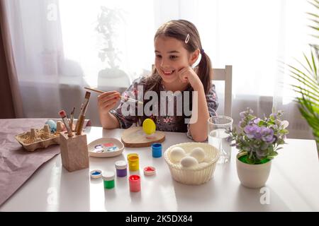 Charmantes kleines Mädchen sitzt an einem weißen Tisch am Fenster, lächelt, hält einen Pinsel, ein bemaltes Ei steht vor ihr. Stockfoto