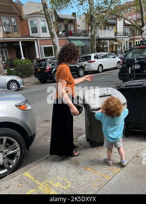 Das kleine Mädchen hilft Mama, den Müll zur Abholung der sanitären Anlagen in einer Wohnstraße in Brooklyn, New York, zu entsorgen. Stockfoto