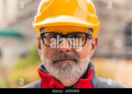 Nahaufnahme Porträt eines ernsten Gesichts des kaukasischen Mannes mit grauem Gesichtshaar und einer Brille mit schwarzem Rahmen, die einen gelben Schutzhelm trägt. Engineering und Architektur. Hochwertige Fotos Stockfoto