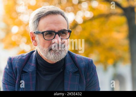 Nahaufnahme des kaukasischen Mannes im Jahr 60s mit grauem Haar und grauem Bart, Brille mit schwarzem Rahmen, Blazer und schwarzem Rollkragen, Blick auf etwas außerhalb der Aufnahme und leicht lächelnd. Großer Baum mit orangefarbenen Blättern im verschwommenen Hintergrund. Entspannung im Herbst im Freien. Hochwertige Fotos Stockfoto