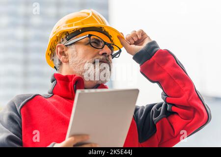 Erfahrener professioneller Architekt, der die laufenden Arbeiten mit seinem Projekt auf einem tragbaren Tablet-PC unternahm, während er seinen gelben Helm mit einer Hand hielt. Schwarz-rotes Fleece. Außenportrait. Hochwertige Fotos Stockfoto