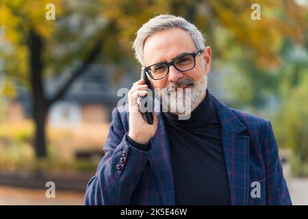 Hübscher männlicher grauhaariger kaukasischer Firmenbesitzer, der sein schwarzes Smartphone in der Nähe seines Ohrs hält und einen Anruf entgegenruft. Verschwommene Parkbäume im Hintergrund. Karierter Blazer und Rollkragen. Hochwertige Fotos Stockfoto