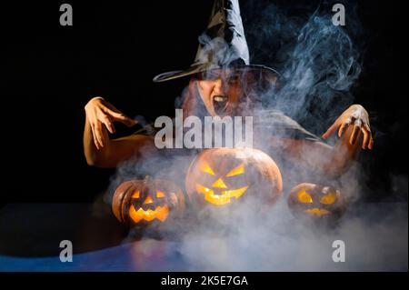 Eine ominöse Hexe im Hut zaubert über eine Jack-o-Laterne. Traditionelle halloween-Figuren. Mystischer Nebel kriecht über Kürbisse mit geschnitzten schrecklich Stockfoto